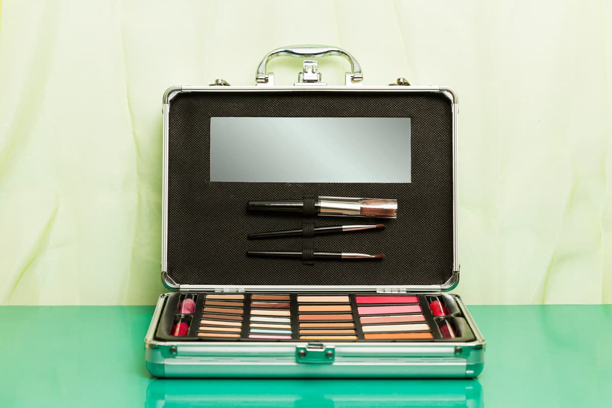 A silver aluminum open makeup case with eye shadows, blushes, pencils and brushes on a green background. 