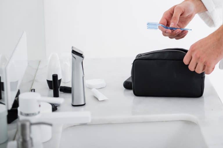 man hands holding a blue toothbrush inside a white bathroom, holding black toiletry bag, clean white bathroom sink, toiletry stuffs, Do Toiletry Bags Need To Be Clear?