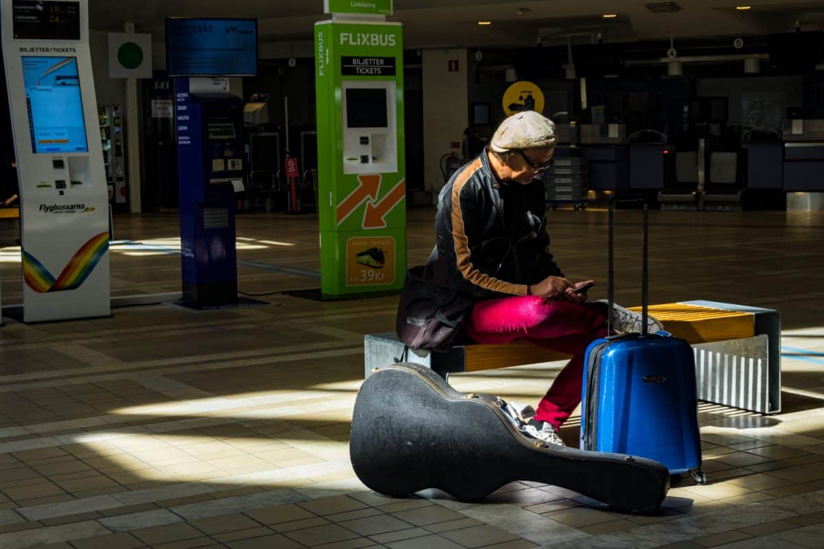A man sits on a bech at Arlanda airport with a guitar case and a suitcase.