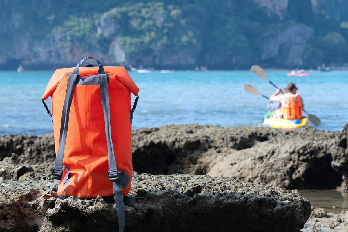 Get a dry bag - Vacation in Thailand. Big orange waterproof backpack (dry bag) on the blurry background with sea and people in a kayak.
