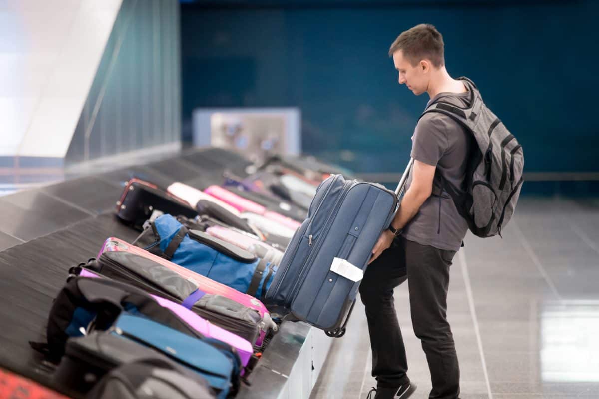 Young handsome man passenger in 20s with carry-on backpack collecting his luggage at conveyor belt in arrivals lounge of airport terminal building

