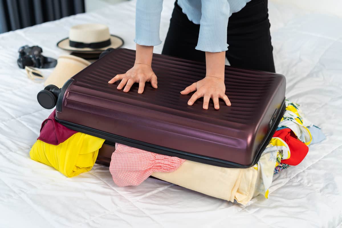 Woman trying to close overfilled suitcase on her bed.