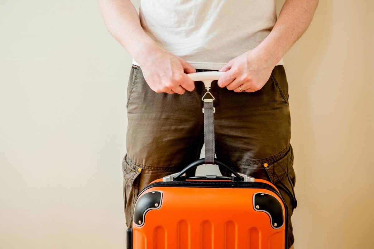 Man tourist using digital luggage scale at home to weighs luggage to avoid overweight baggage in airport concept. Reduce traveling stress.
